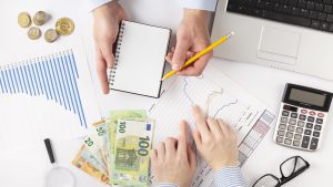 Rokas ar naudu, kalkulatoru un grafikiem, kas simbolizē finanšu plānošanu