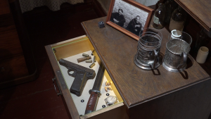 Atvērta naktsgaldiņa atvilktne ar pistoli, munīciju, nazi, pulksteni un citiem priekšmetiem, blakus atrodas divas tējas glāzes un veca fotogrāfija.