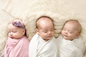 Trīs zīdaiņi, cieši ietīti siltos pārvalkos un guļot līdzās, viens no viņiem rotāts ar violeto galvas lentu