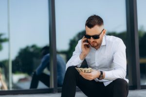 Vīrietis baltā kreklā ar saulesbrillēm skaita naudu un runā pa mobilo tālruni