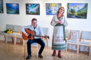 Sieviete latviešu tradicionālā tērpā dzied, kamēr vīrietis blakus spēlē akustisko ģitāru mākslas darbu izstādes zālē