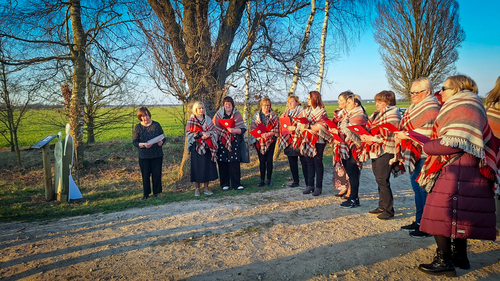 Sieviešu dziedātāju grupa tautiskās segās dzied pie piemiņas zīmes laukā