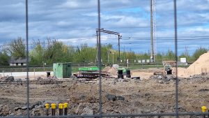 Būvniecības laukums ar smilšu kaudzi, zaļiem konteineriem un elektroinstalācijām