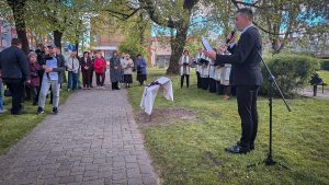 Vīrietis runā mikrofonā pie pieminekļa atklāšanas ceremonijas, apkārt stāv cilvēki