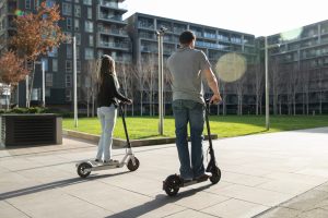 Divi cilvēki, viens vīrietis un viena sieviete, brauc ar elektriskajiem skrejriteņiem modernā pilsētas kvartālā