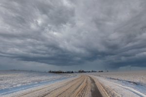 Lauku ceļš ziemā ar smagiem vētras mākoņiem debesīs, tālumā redzams neliels ciemats.