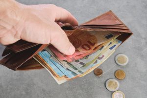 Roka tur atvērtu brūnu ādas maciņu ar eiro banknotēm, bet blakus uz galda redzamas vairākas eiro monētas