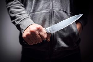 Persona pelēkā džemperī cieši tur rokā asu nazi, radot draudīgu situāciju