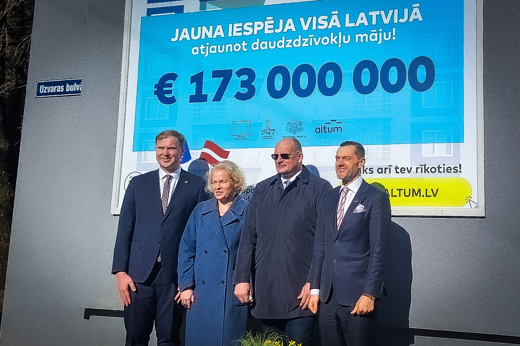 Četri cilvēki stāv pie liela plakāta, kur redzams 173 miljonu eiro atbalsts daudzdzīvokļu māju