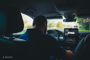 Automašīnas salonā redzams cilvēks pie stūres, priekšējā panelī uzstādīts policijas aprīkojums ar ekrānu