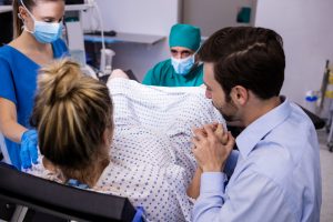 Topošā mamma dzemdību gultā ar partneri blakus, dzemdību personālam asistējot un valkājot sejas maskas