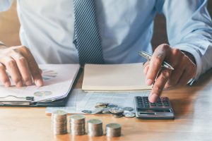 Biznesa cilvēks uzvalkā ar kalkulatoru un monētām uz galda, aprēķinot finanses