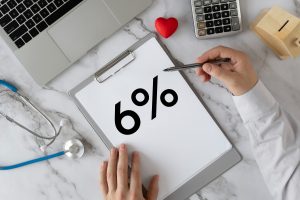 Dokumentā uz rakstāmgalda redzams uzraksts “6%”, apkārt klēpjdators, stetoskops un kalkulators.