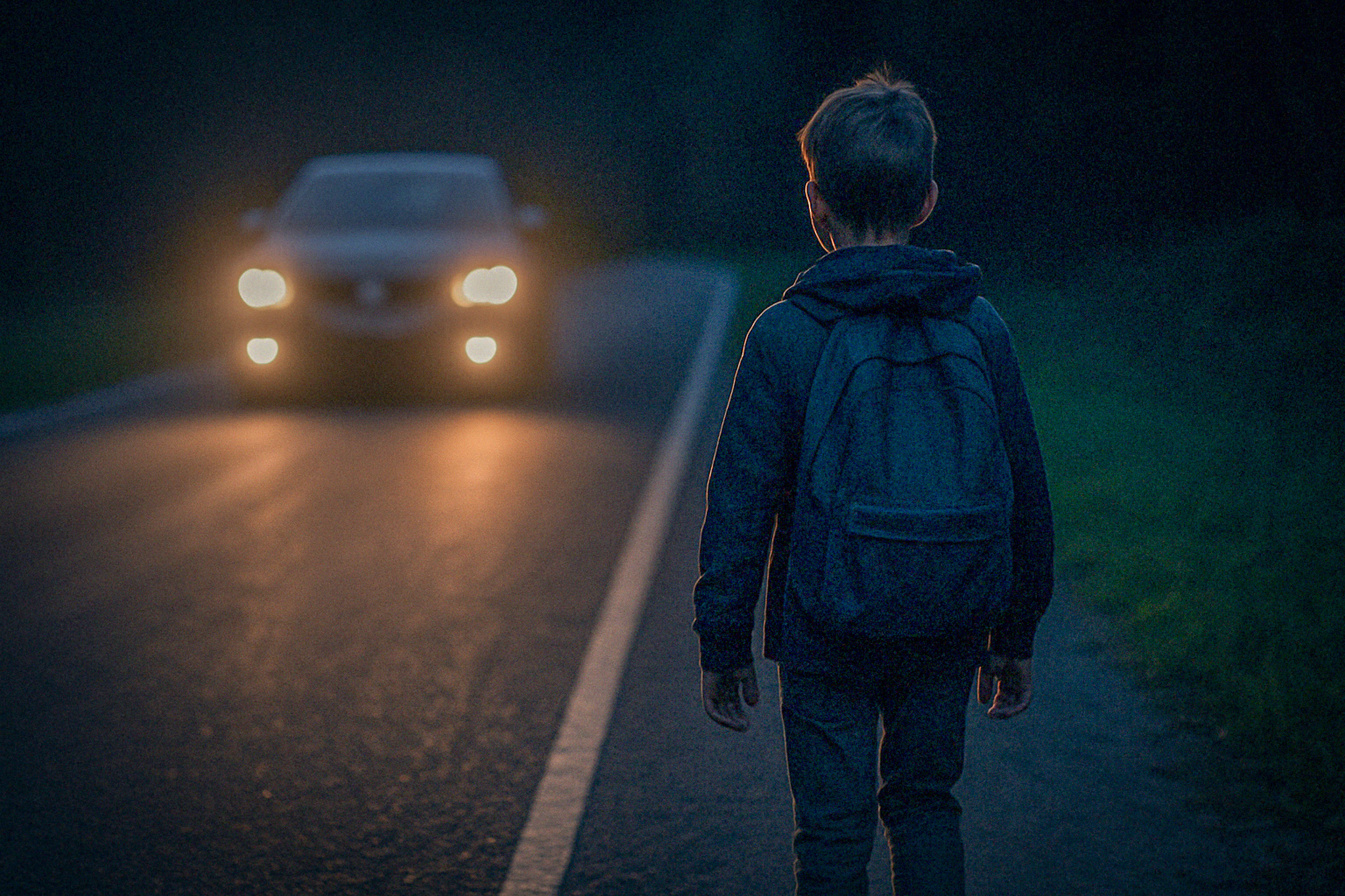 Zēns ar mugursomu tumsā uz ceļa, pretim tuvojas automašīna ar ieslēgtiem lukturiem