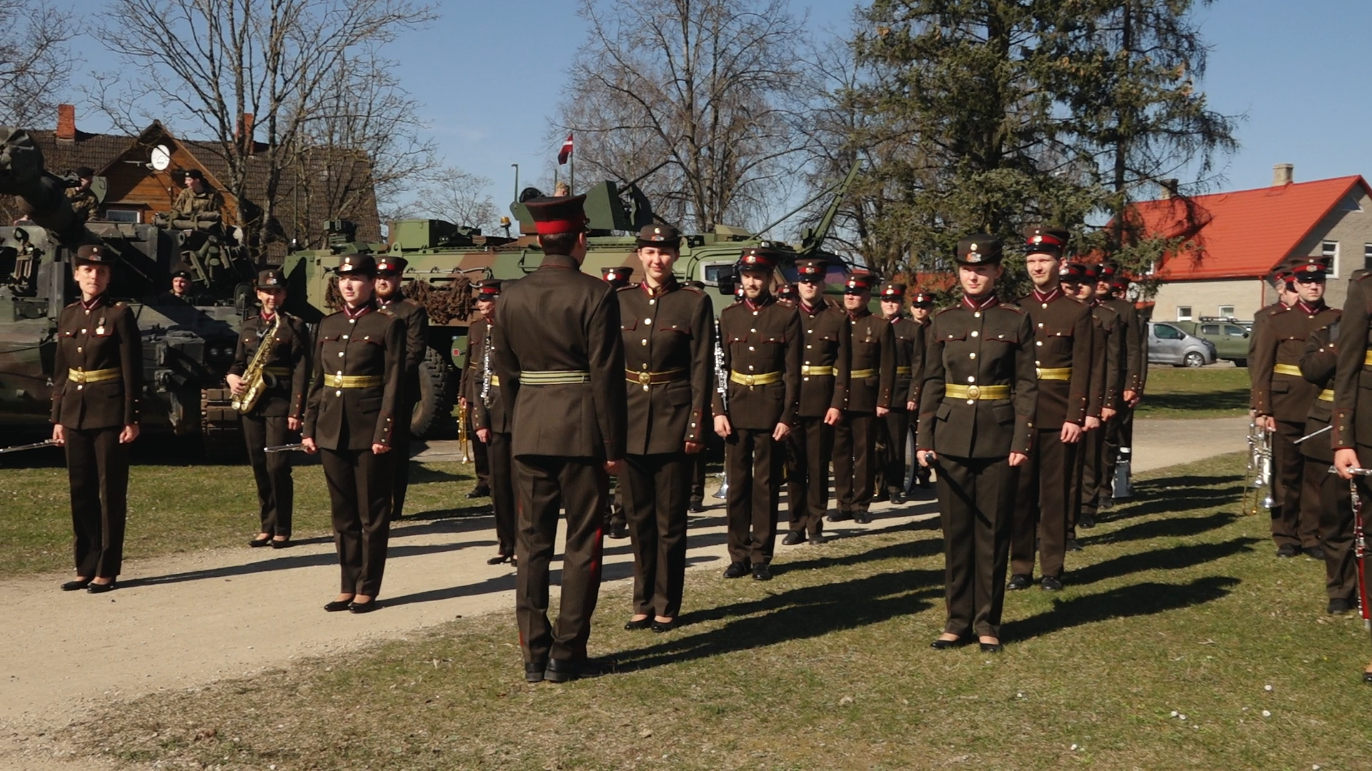 Militārās formas tērptu karavīru ierinda ceremonijā ar bruņutehniku fonā