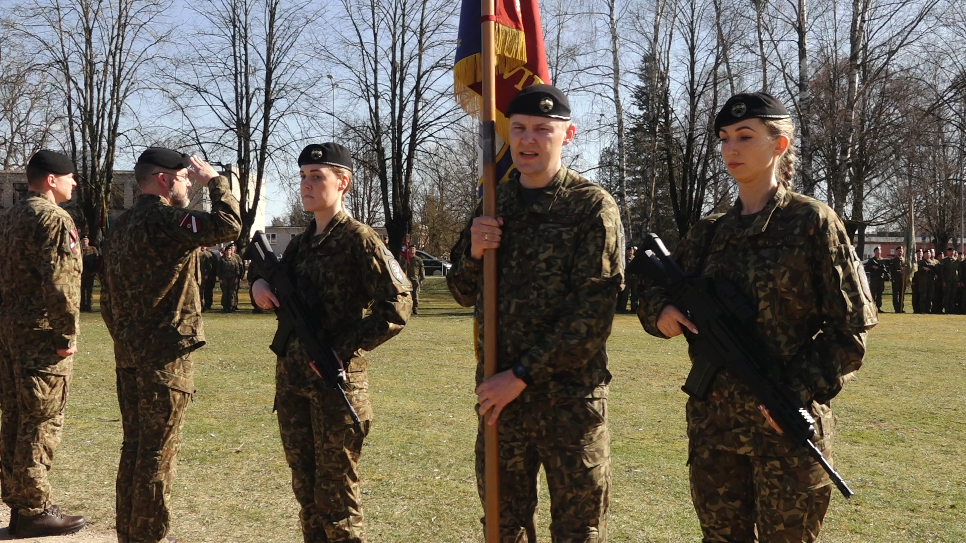 Latvijas karavīri kamuflāžas formās ar ieročiem un vienības karogu militārā ceremonijā.