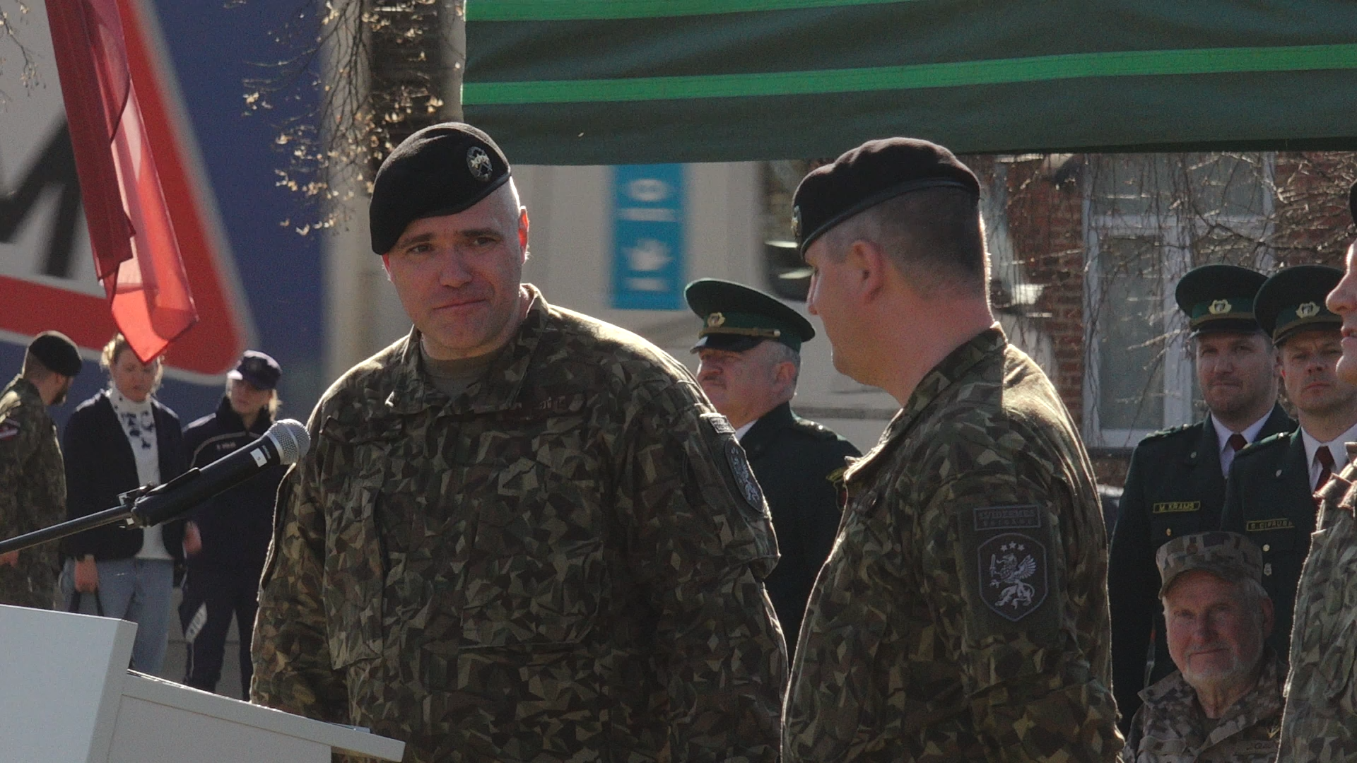 Militārpersonas kamuflāžas formās runā pie mikrofona ceremonijas laikā, fonā redzami robežsargi un skatītāji.