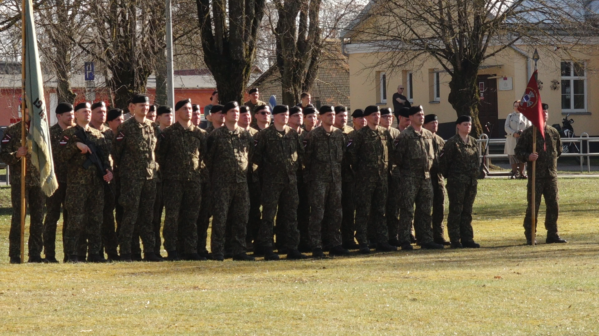 Latvijas karavīru vienība kamuflāžas formās stāv ierindā ar karogiem militārā ceremonijā.