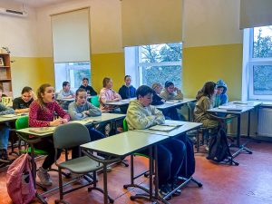 Skolēni sēž klasē pie galdiem, klausoties mācību stundā un pildot uzdevumus