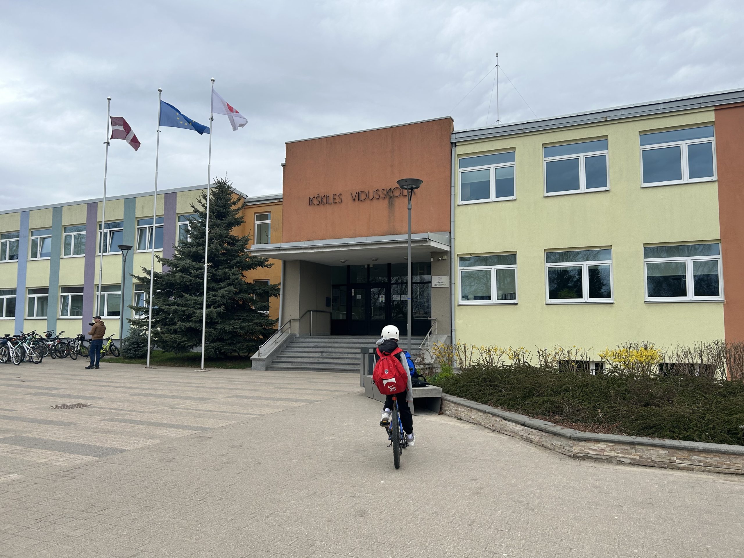 Skolēns ar sarkanu mugursomu tuvojas Ikšķiles vidusskolas ēkai ar velosipēdu
