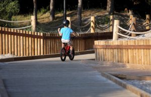 Bērns ar ķiveri brauc ar velosipēdu pa koka taku piejūras zonā