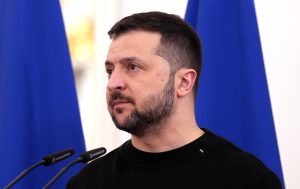 Volodimirs Zelenskis melnā džemperī runā pie mikrofoniem ar ziliem fonā esošām karogiem
