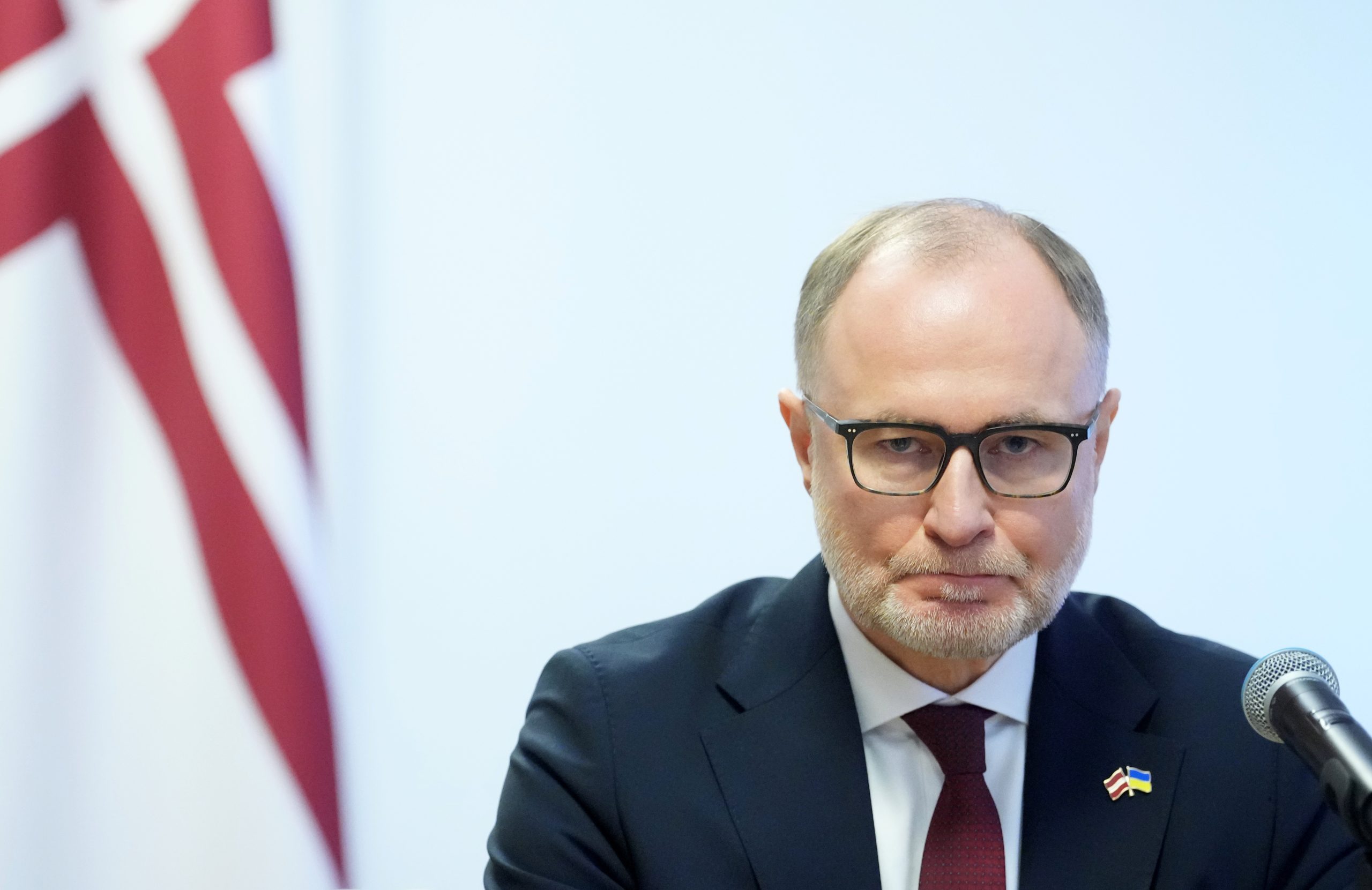 Andris Sprūds uzvalkā ar brillēm preses konferencē pie mikrofona, fonā Latvijas karogs