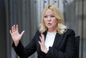 Sieviete ar blondiem matiem un melnu žaketi žestikulē, runājot publiskā uzstāšanās brīdī