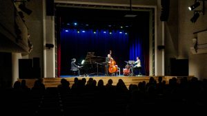 Koncerts uz skatuves, kur muzicē pianists, kontrabasists un sitaminstrumentālists.