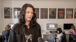Jaunietis Lauris Bogdanovičs ar gariem, tumšiem matiem intervijas laikā, fonā redzami datori un plakāti radošajā klasē