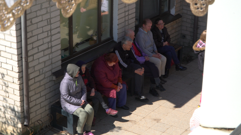 Gados vecāki cilvēki sēž saulē pie ēkas sienas