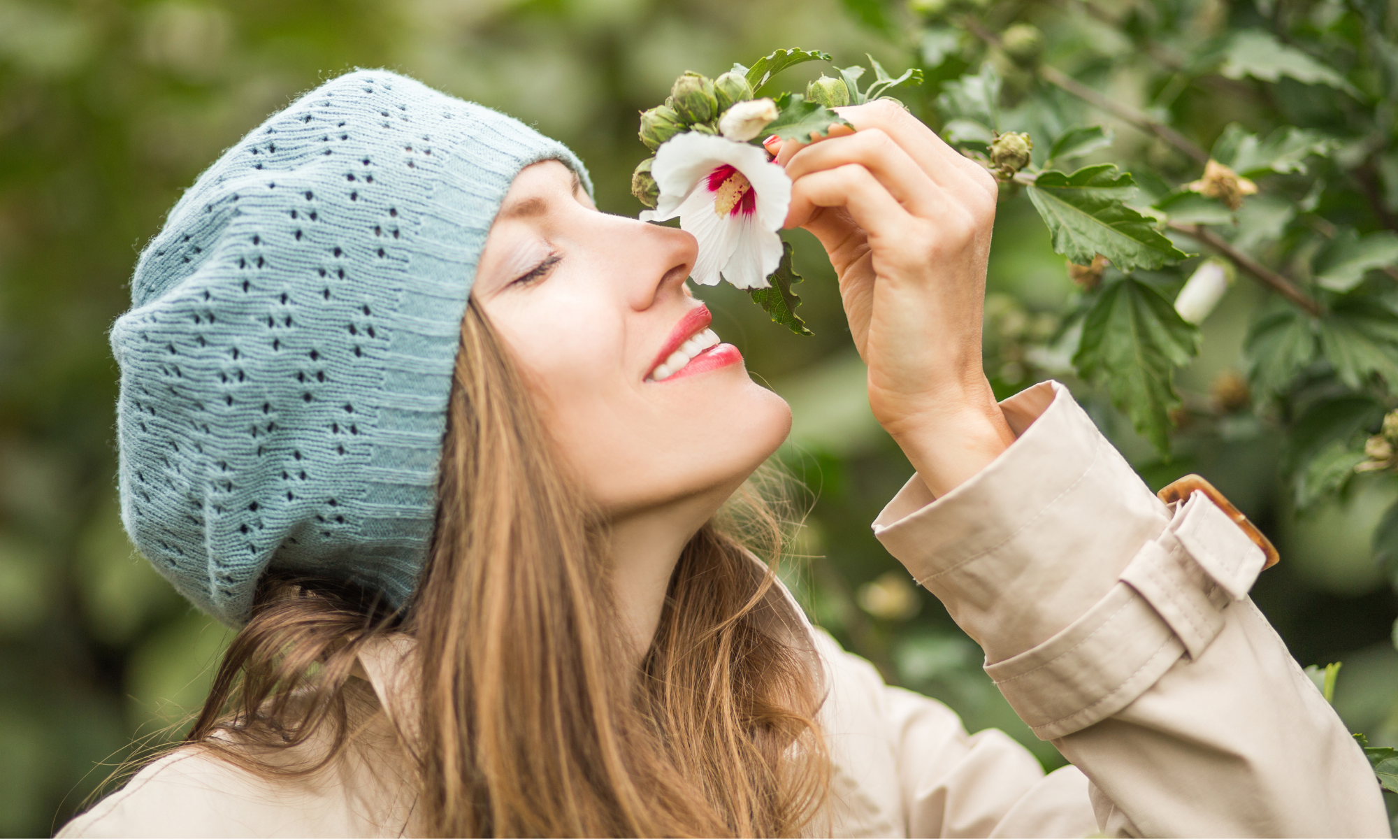 Sieviete ar zilu adītu cepuri smaida un smaržo ziedus hibiska ziedu dārzā