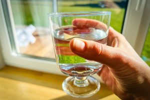 Roka tur nelielu glāzi ar dzeramo ūdeni, fonā redzama zaļa ainava aiz loga
