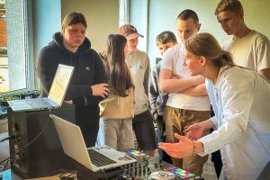 Jaunieši pulcējušies ap DJ pulti un portatīvajiem datoriem, kamēr instruktore skaidro mūzikas miksēšanas principus
