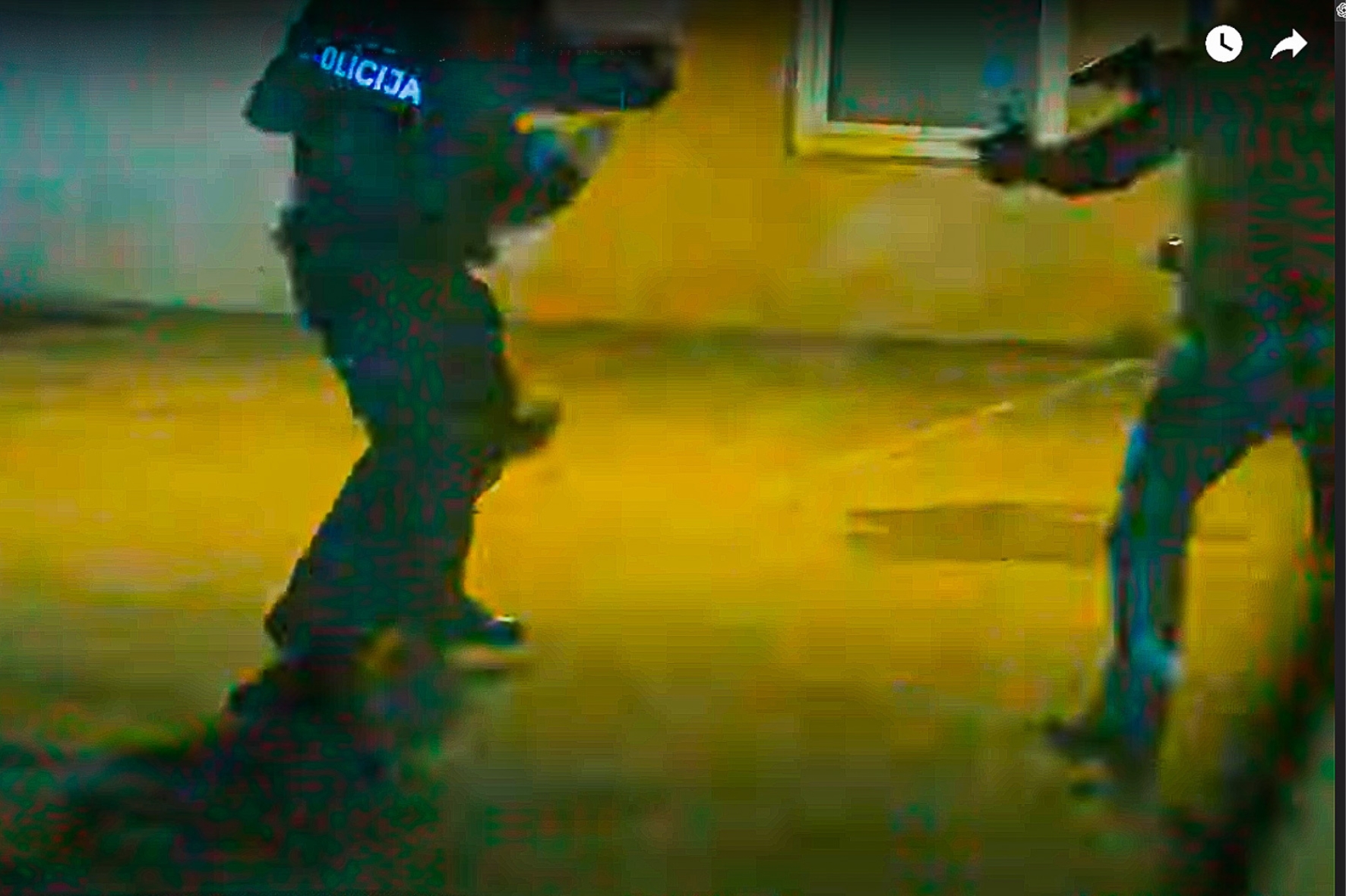 Policijas formā tērpts cilvēks, iespējams, iesaistīts fiziskā konfliktā, dzeltenīgi izgaismotā pilsētvidē