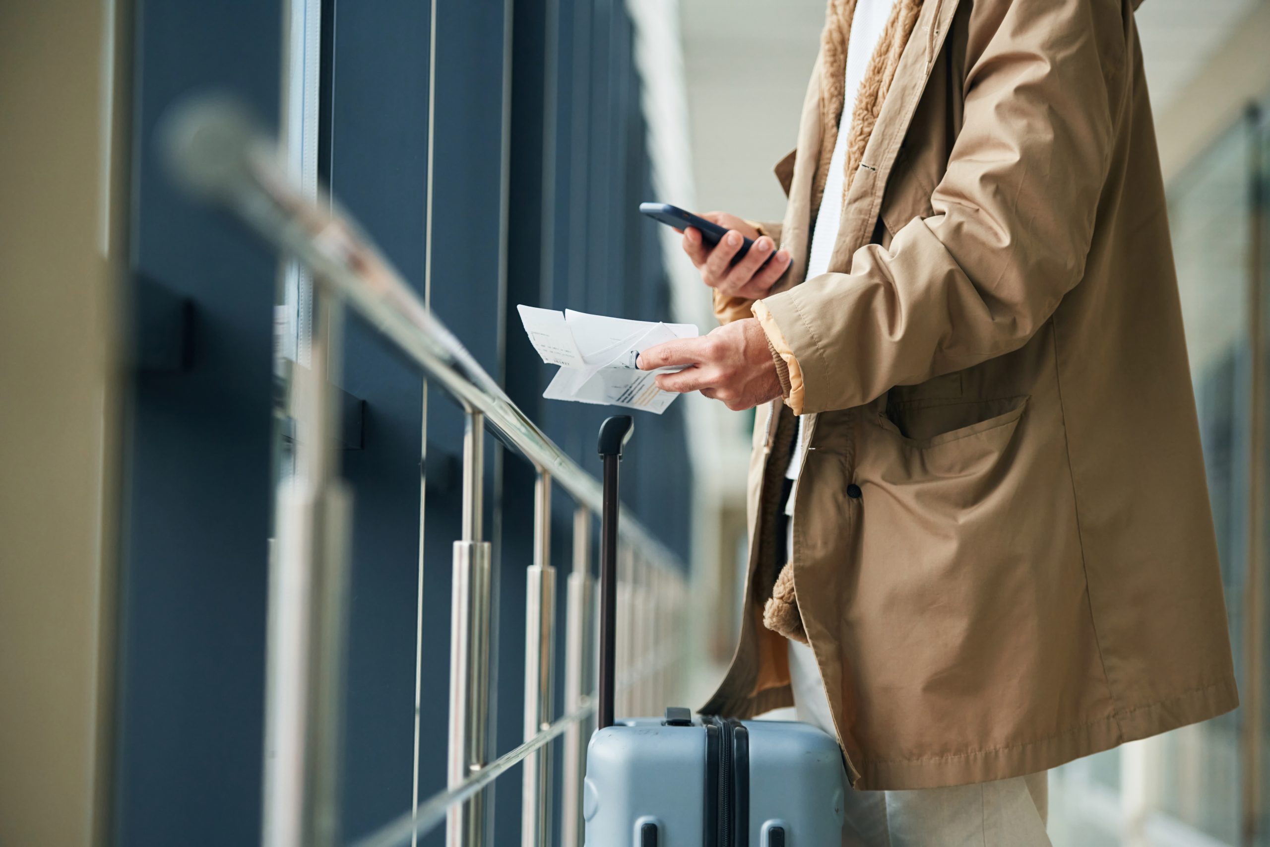 Vīrietis ar čemodānu gaida lidostā, skatoties mobilajā telefonā.