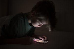 Bērns tumsā lieto viedtālruni, skatoties sociālo tīklu saturu