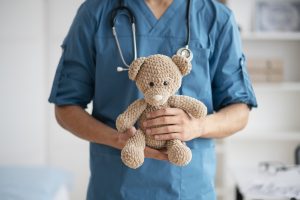 Ārsts tērpies zilā darba apģērbā tur adīto lāču rotaļlietu bērnu pediatrijas birojā
