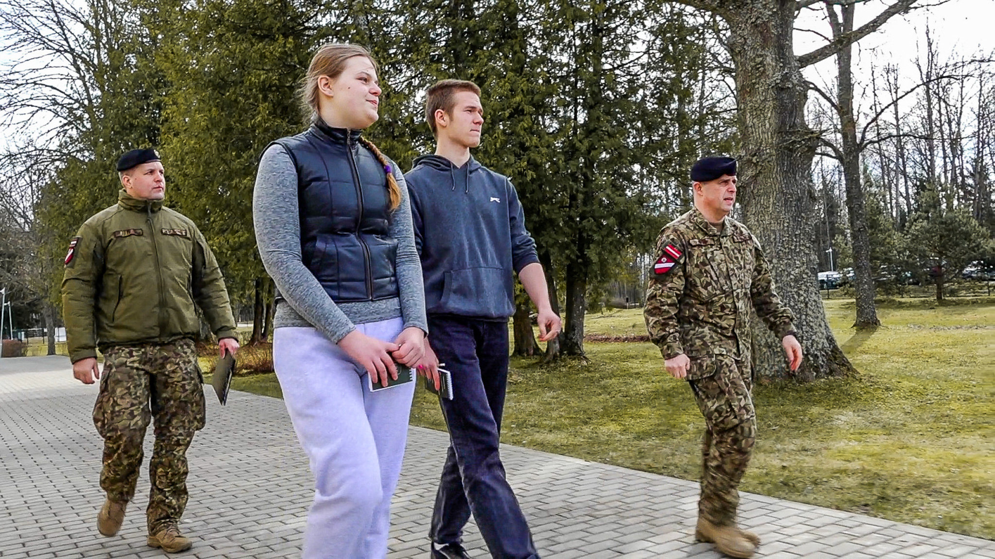 Divi jauni civiliedzīvotāji un divi militārā formā tērpti instruktori pastaigā pa bruģētu celiņu