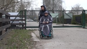 Sieviete stumj ratiņkrēslā sēdošu senioru pie slēgtiem vārtiem pilsētvidē