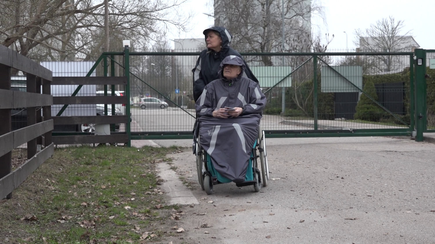 Sieviete stumj ratiņkrēslā sēdošu senioru pie slēgtiem vārtiem pilsētvidē