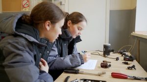 Divas meitenes strādā tehniskajā darbnīcā, veicot pierakstus un pētot mehāniskās detaļas