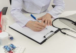Ārste baltā halātā raksta pierakstus uz klipša dēļa pie galda ar medicīnas piederumiem.