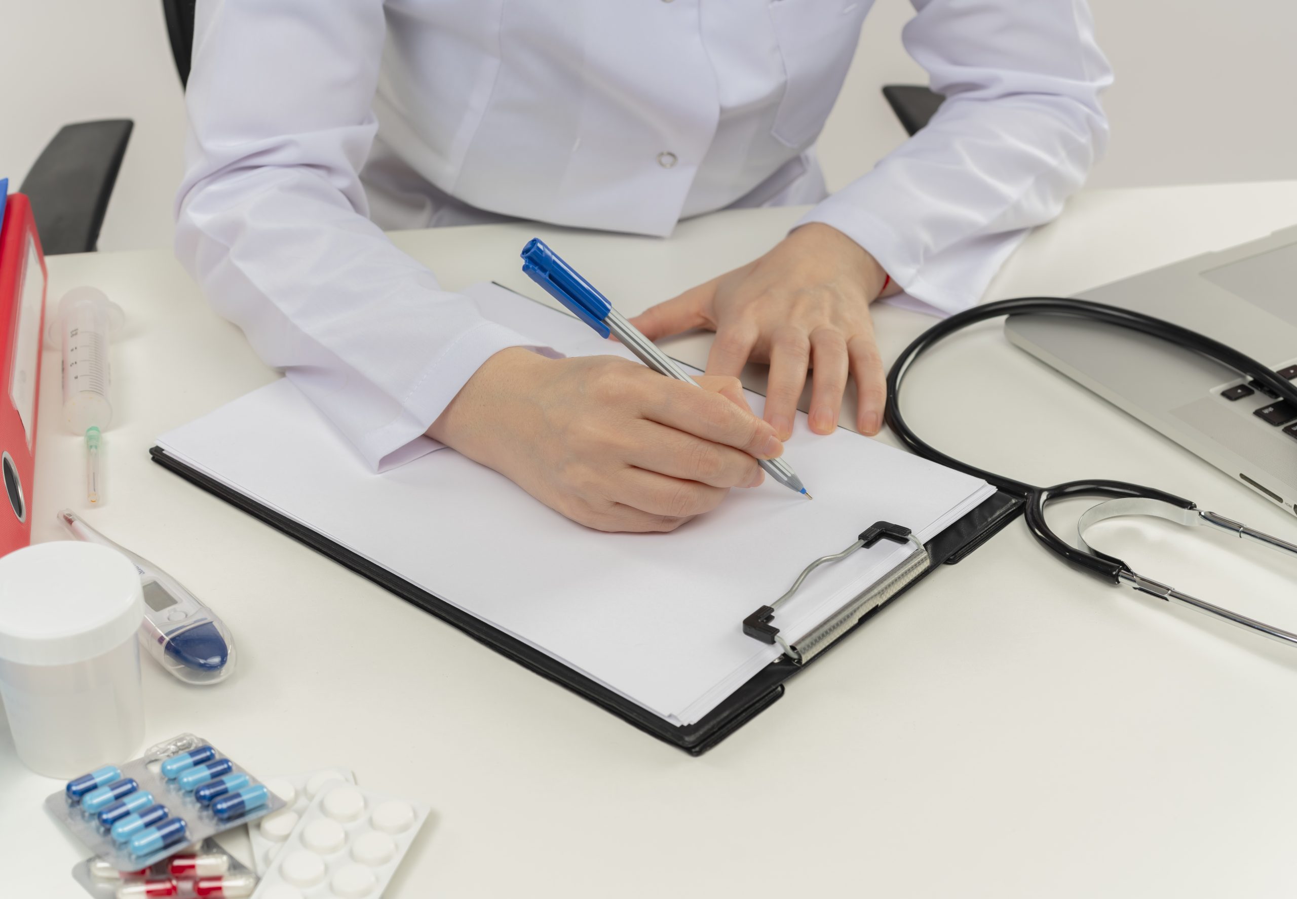 Ārste baltā halātā raksta pierakstus uz klipša dēļa pie galda ar medicīnas piederumiem.
