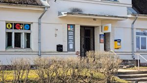 Pasta ēkas ieeja ar pakalpojumu norādēm uz fasādes un dzeltenu pastkastīti pie sienas