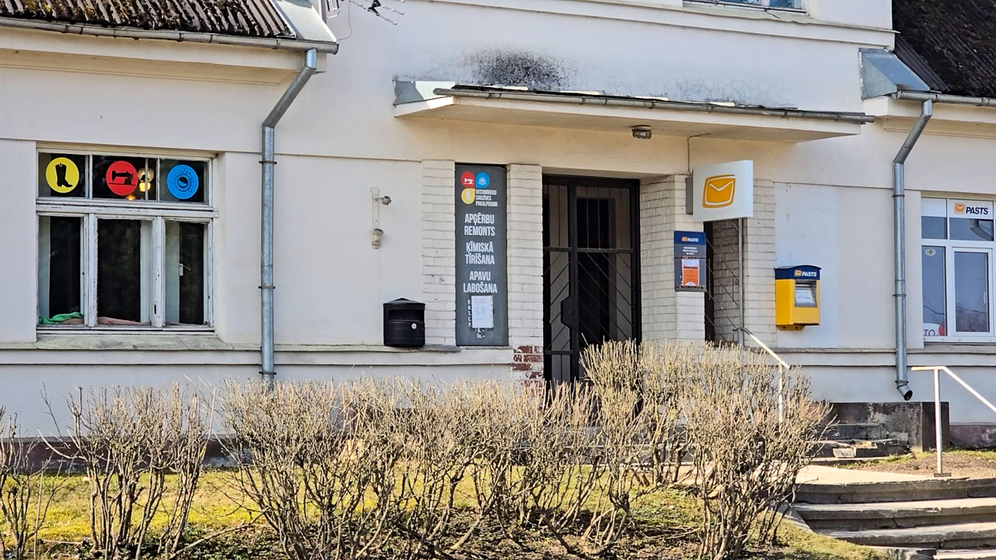 Pasta ēkas ieeja ar pakalpojumu norādēm uz fasādes un dzeltenu pastkastīti pie sienas