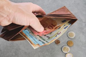 Rokā turēts maks ar eiro banknotēm un monētām uz galda.