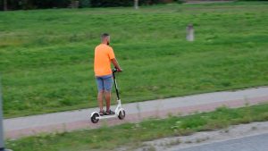 Vīrietis ar elektrisko skrejriteni pārvietojas pa ietvi zaļā pilsētvidē.