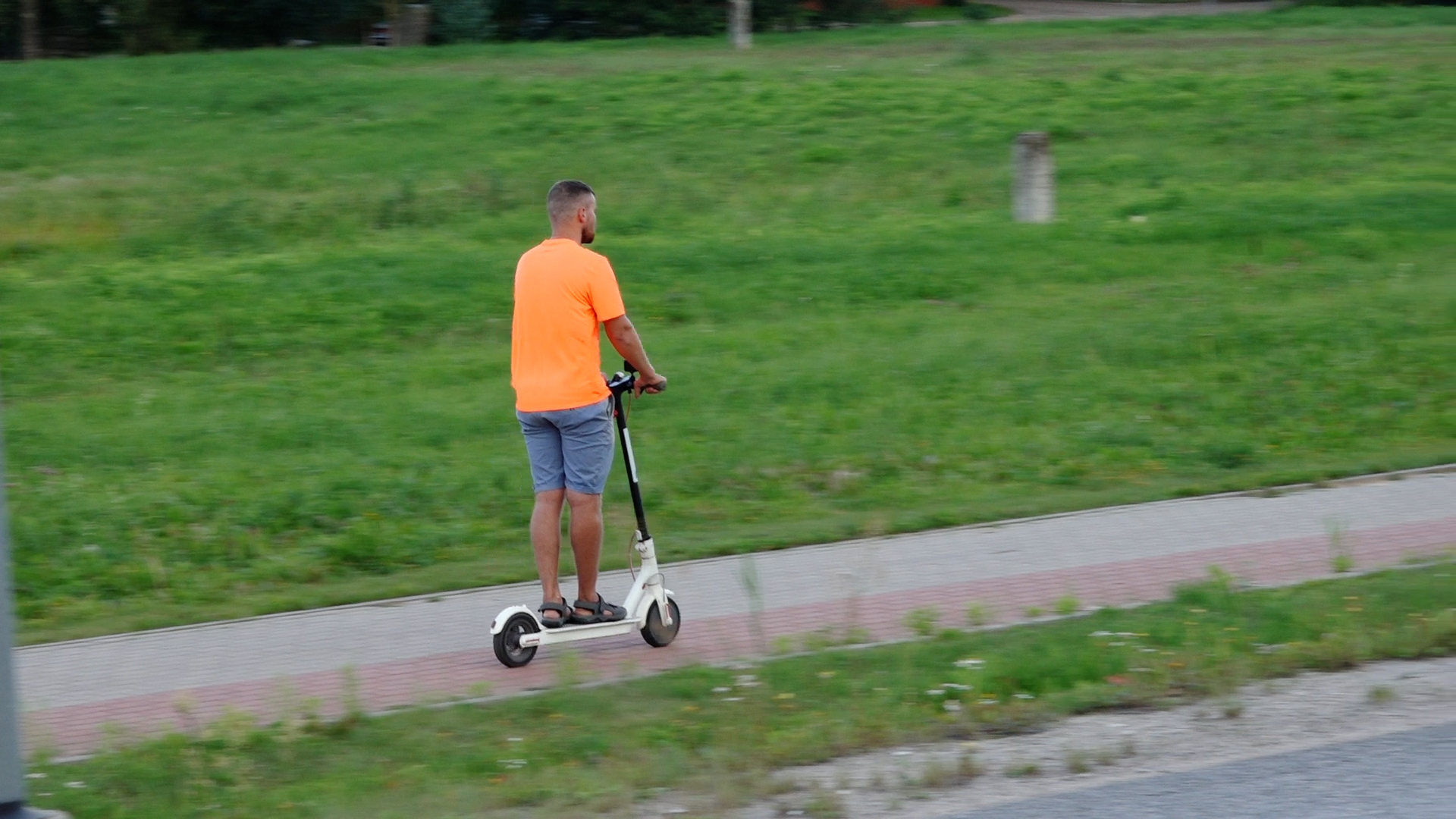 Vīrietis ar elektrisko skrejriteni pārvietojas pa ietvi zaļā pilsētvidē.