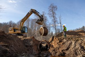 Ekskavators uzstāda caurteku upē – ceļu būvniecības darbi dabā